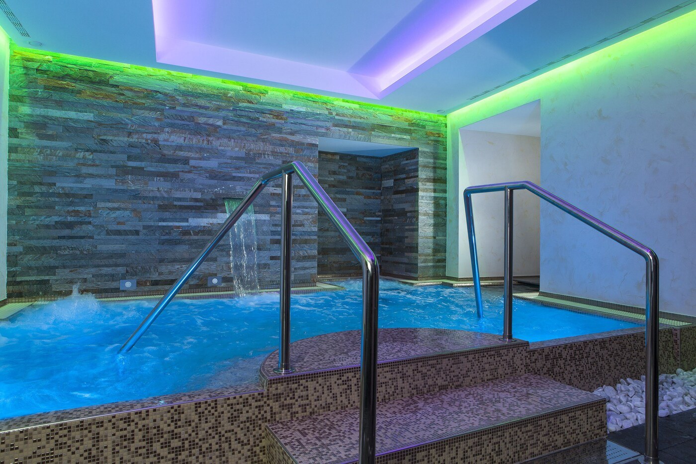 A hot tub with steps and metal railings, surrounded by mosaic-tiled walls and a stone-like textured wall. - Hotel Artemide (Rome)