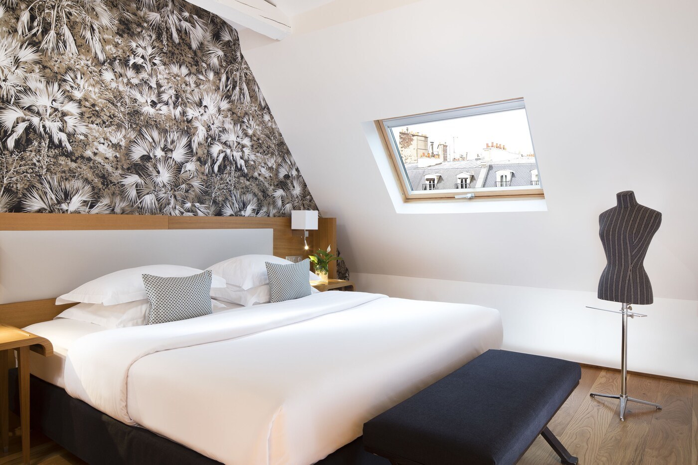 A hotel room with a large bed, a bench, a mannequin, a window with a city view, and a wall with a leaf pattern. - Hotel Le Six (Paris)