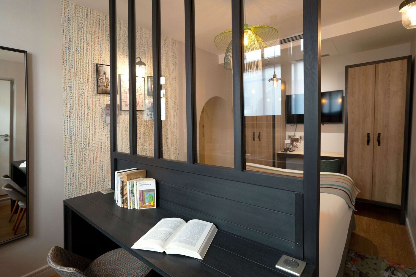 A modern bedroom with a dark wooden headboard, a bedside table with books, a bed with a striped blanket, and a TV mounted on the wall. - Hôtel Littéraire Marcel Aymé, BW Premier Collection (Paris)